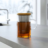 Glass mug with wooden lid containing tea on a kitchen counter.