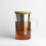 A clear glass mug with a bamboo lid, a stainless steel infuser, and a clear glass handle, filled with amber tea.