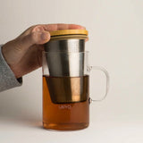 Glass tea infuser with wooden lid held by a hand, Ukiyo branding visible, on a light background.